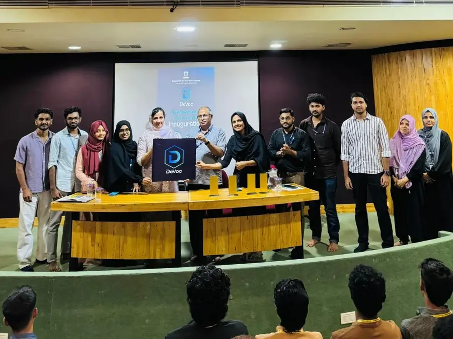 Group photo of the DeVoc team during logo launch on stage with Farook College Principal Dr. Aysha Swapna and department faculty